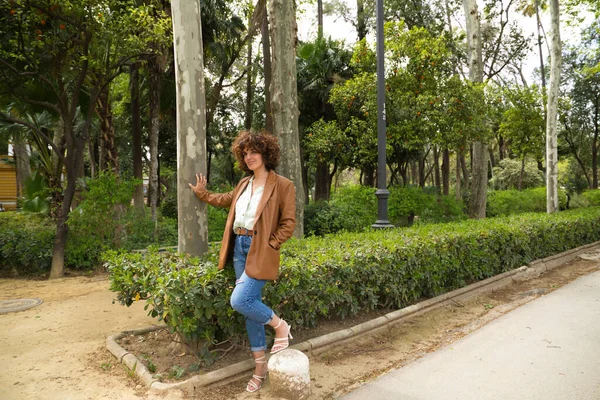 Orta yaşlı, güzel, kıvırcık saçlı İspanyol bir kadın bir Avrupa şehrinde büyük ağaçları olan bir parkta duruyor. Kadın modern ve modaya uygun kıyafetler giyiyor. Seyahat ve tatil konsepti.