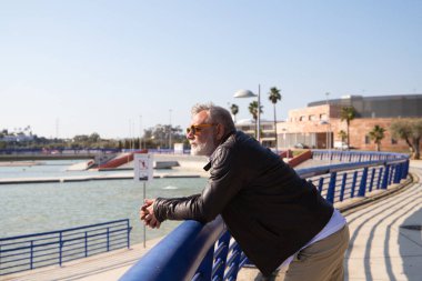 Sakallı ve gri saçlı yakışıklı olgun bir adam parkta yürüyor. Adam bej pantolon, beyaz gömlek ve deri ceket giyiyor. Yaşlılık kavramı