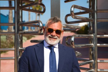 Yakışıklı, olgun, gri saçlı, sakallı, ceketli, kravatlı ve güneş gözlüklü bir adam. Baş muhasebeci ve yönetici olarak çalıştığı ofis binasının girişinde. Olgun iş adamı konsepti