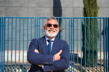Mature, grey-haired, bearded man with sunglasses, jacket and tie makes different expressions for the photo, looks over his sunglasses and touches his lips. Concept various expressions.