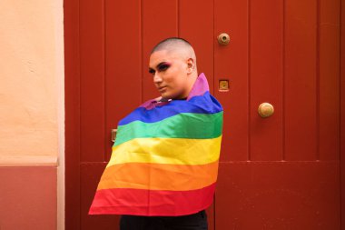 İkili olmayan, Güney Amerika kökenli ve genç biri, makyaj yapıyor ve omuzlarında eşcinsel gururu bayrağı taşıyor. Eşitlik kavramı, homoseksüellik, gay, lezbiyen, gay gururu.