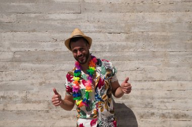 Havai çiçekli gömlek, şapka ve renkli çiçekli kolye giyen yakışıklı bir genç. Adam çok mutlu. Arka plan gri duvar. Seyahat ve tatil konsepti.