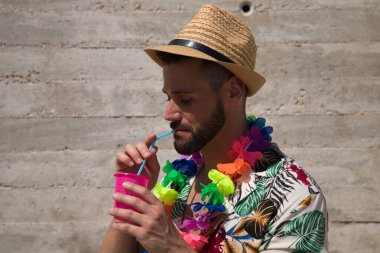 Hawaii çiçekli gömlek, şapka ve renkli çiçek kolyesi giyen yakışıklı genç adam, içinde meşrubat olan bir bardaktan bir kamışdan su içiyor. Tatil ve seyahat kavramı.