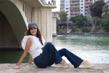 Genç, güzel, siyah, kıvırcık saçlı ve burnu kalkık bir kadın kep takıyor ve nehir kenarında oturuyor. Kadın fotoğraf için poz veriyor. Kavramsal ifadeler. Gülümse, üzül, düşün, eğlen, yaşa.