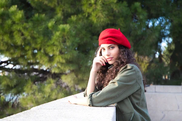 Beautiful young latina woman with curly brown hair wearing a red cap and dressed in casual clothes is sightseeing in Europe. She is posing for photos. Mediterranean pine tree in the background.Tourism