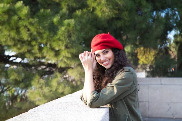 Beautiful young latina woman with curly brown hair wearing a red cap and dressed in casual clothes is sightseeing in Europe. She is posing for photos. Mediterranean pine tree in the background.Tourism