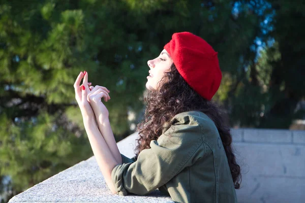 Beautiful young latina woman with curly brown hair wearing a red cap and dressed in casual clothes is sightseeing in Europe. She is looking at the horizon. Mediterranean pine tree in the background.