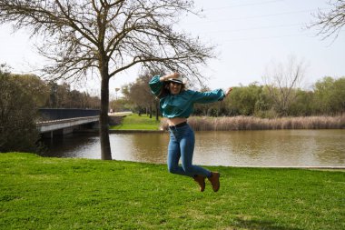 Güzel, sarışın, genç bir kadın elinde şapkasıyla nehir kıyısında zıplıyor. Kadın çok mutlu. Konsept ifade ve rahatlama süresi.