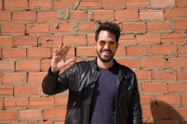 Genç Latin bir adam, esmer, sakallı ve el kaldırarak vücut bulmuş. Arka planda kırmızı tuğla duvar var. Merhaba de, güle güle, selamla. İnsan ilişkileri ve duyguları.