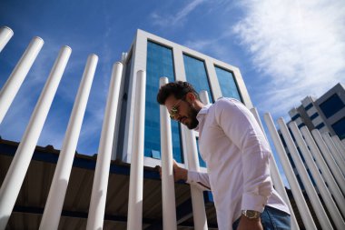 Sakallı, vücudu yontulmuş ve güneş gözlüklü yakışıklı genç adam beyaz çitlere tutunuyor. Fotoğraf aşağıdan çekildi ve arka planda mavi gökyüzünü görebilirsiniz..