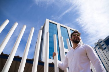 Sakallı, vücudu yontulmuş ve güneş gözlüklü yakışıklı genç adam beyaz çitlere tutunuyor. Fotoğraf aşağıdan çekildi ve arka planda mavi gökyüzünü görebilirsiniz..