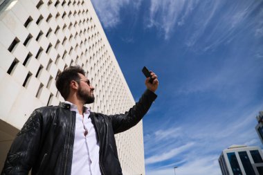 Sakallı, heykel vücutlu yakışıklı genç adam sokakta cep telefonuyla selfie çekiyor..