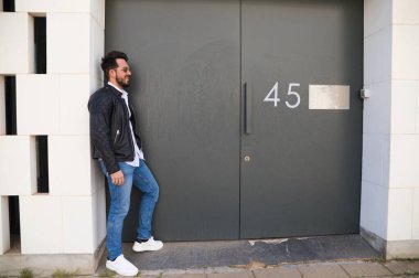 Sakallı, vücudu yontulmuş ve güneş gözlüklü yakışıklı genç adam portre için farklı pozlar verirken garajının gri kapısında duruyor. Farklı ifadeleri kavra.