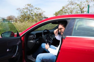 Genç yakışıklı bir adam, kırmızı spor arabasında oturan heykel gibi vücudu var. Adam zengin ve modern kıyafetler giymiş. Yüksek yaşam standardı ve finansal olarak iyi konumlandırılmış..