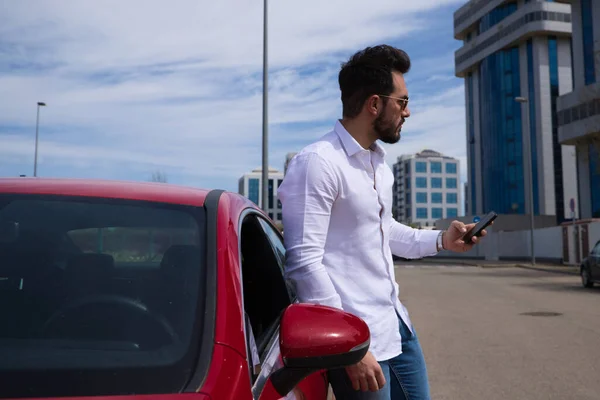 Handsome young man, looking at his mobile phone next to his red sports car. The man is wealthy. High standard of living and well positioned financially.