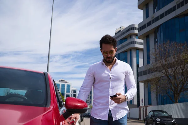Handsome young man, looking at his mobile phone next to his red sports car. The man is wealthy. High standard of living and well positioned financially.