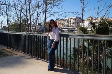 young and beautiful woman from south america touring europe. The woman is wearing a red flower in her hair and is leaning on the railing next to the river in sevilla. Travel and tourism concept.