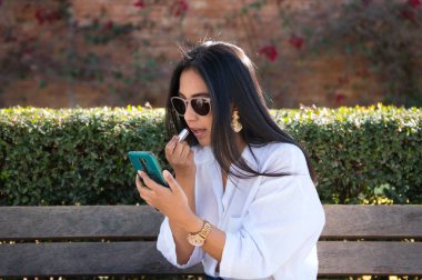 Avrupa 'da tatilde olan genç ve güzel bir Güney Amerikalı kadın cep telefonunu ayna olarak kullanarak bir bankta dudaklarını boyuyor..