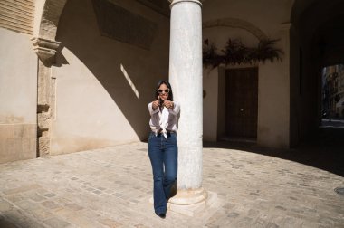 Genç, güzel, esmer, Güney Amerikalı kadın Avrupa 'da seyahat ediyor ve beyaz mermer bir sütuna yaslanmış ellerini kaldırarak baş parmağını kaldırıyor. Seyahat ve tatil konsepti.