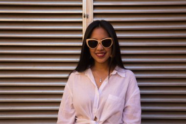 Genç, güzel, siyah saçlı, beyaz gömlek ve güneş gözlüğü takan Güney Amerikalı kadının arka planda gümüş renkli bir hava akışı var. Kadın farklı ifadeler kullanıyor..