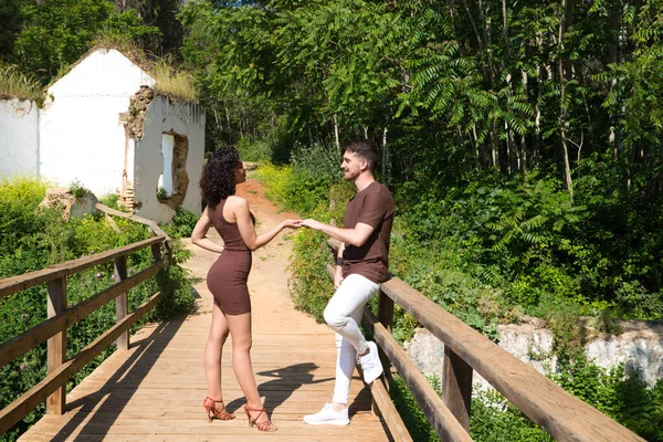 Young and handsome latin dance couple salsa and bachata in the park on a wooden bridge. The couple dances passionately and in love surrounded by greenery and trees. Dance concept.