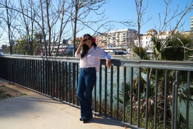 young and beautiful woman from south america touring europe. The woman is wearing a red flower in her hair and is leaning on the railing next to the river in sevilla. Travel and tourism concept.
