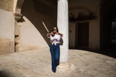 Genç, güzel ve esmer Güney Amerikalı kadın Avrupa 'da seyahat ediyor ve elleriyle bir kalp oluştururken beyaz mermer bir sütuna yaslanıyor. Seyahat ve tatil konsepti.