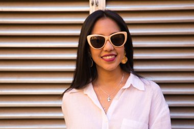 Genç, güzel, siyah saçlı, beyaz gömlek ve güneş gözlüğü takan Güney Amerikalı kadının arka planda gümüş renkli bir hava akışı var. Kadın farklı ifadeler kullanıyor..