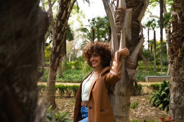 Orta yaşlı, güzel, kıvırcık saçlı İspanyol bir kadın bir Avrupa şehrinde büyük ağaçları olan bir parkta duruyor. Kadın modern ve modaya uygun kıyafetler giyiyor. Seyahat ve tatil konsepti.