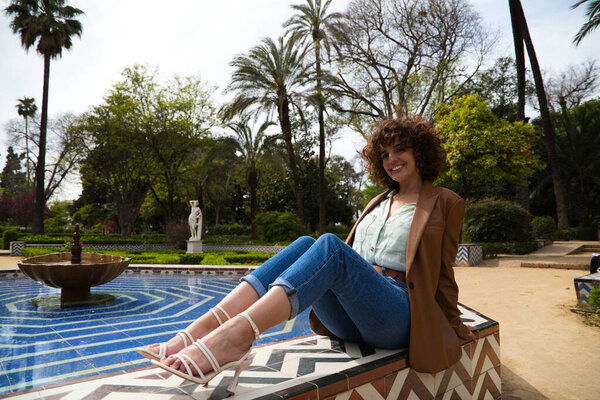 красивая женщина с вьющимися волосами, прислонившаяся к фонтану в средиземноморском и мусульманском стиле в парке в Севилье. Женщина одета в современную одежду. Концепция путешествия и отдыха, отдых и релаксация.