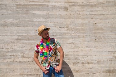 Havai çiçekli gömlek, şapka ve renkli çiçekli kolye giyen yakışıklı bir genç. Adam çok mutlu. Arka plan gri duvar. Seyahat ve tatil konsepti.