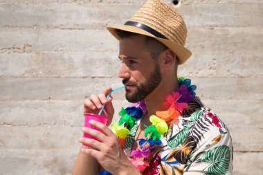 Hawaii çiçekli gömlek, şapka ve renkli çiçek kolyesi giyen yakışıklı genç adam, içinde meşrubat olan bir bardaktan bir kamışdan su içiyor. Tatil ve seyahat kavramı.