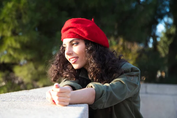 Beautiful young latina woman with curly brown hair wearing a red cap and dressed in casual clothes is sightseeing in Europe. She is looking at the horizon. Mediterranean pine tree in the background.