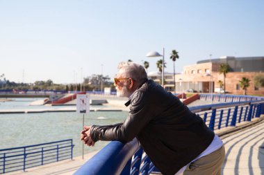 Sakallı ve gri saçlı yakışıklı olgun bir adam parkta yürüyor. Adam bej pantolon, beyaz gömlek ve deri ceket giyiyor. Yaşlılık kavramı