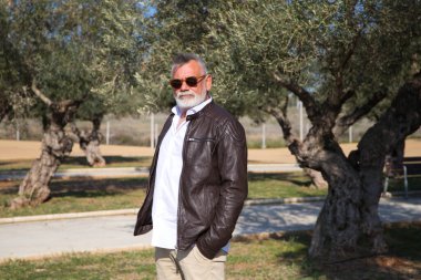 Sakallı ve gri saçlı yakışıklı olgun bir adam parkta yürüyor. Adam bej pantolon, beyaz gömlek ve deri ceket giyiyor. Yaşlılık kavramı
