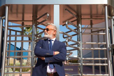 Yakışıklı, olgun, gri saçlı, sakallı, ceketli, kravatlı ve güneş gözlüklü bir adam. Baş muhasebeci ve yönetici olarak çalıştığı ofis binasının girişinde. Olgun iş adamı konsepti