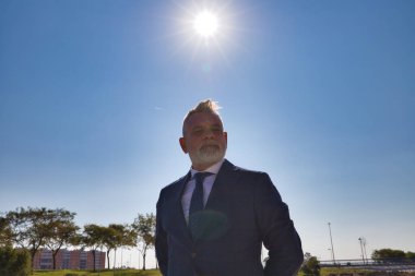 Yakışıklı olgun bir adam, yönetici, gri saçlı, sakallı, ceketli ve kravatlı, arka planda güneş olan bir parkta poz veriyor. Olgun adam, büyükbaba, yönetici kavramı.