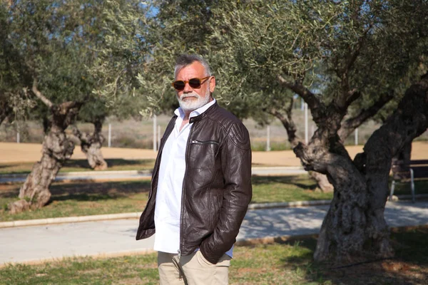 Sakallı ve gri saçlı yakışıklı olgun bir adam parkta yürüyor. Adam bej pantolon, beyaz gömlek ve deri ceket giyiyor. Yaşlılık kavramı