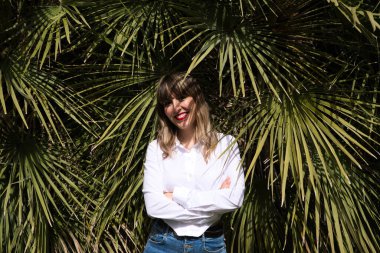 Güzel, sarışın, genç bir kadın parkta yeşil palmiye ağaçlarının önünde farklı ifadeler takınarak eğleniyor. Tatil ve boş zaman kavramı. Çeşitli ifadeler.