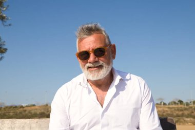 Sakallı ve gri saçlı yakışıklı olgun bir adam kameraya bakarken bir bankta dalgın dalgın oturuyor. Adam bej pantolon ve beyaz gömlek giyiyordu. Yaşlılık ve teknoloji kavramı.