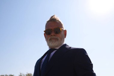 Yakışıklı olgun bir adam, yönetici, gri saç, sakal, güneş gözlüğü, ceket ve kravat, parkta fotoğraf için suni bir göl ile poz veriyor. Olgun adam, büyükbaba, yönetici kavramı.