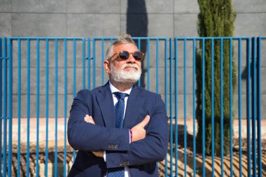 Yakışıklı, olgun, gri saçlı, sakallı, ceketli, kravatlı ve güneş gözlüklü bir adam. Baş muhasebeci ve yönetici olarak çalıştığı ofis binasının girişinde. Olgun iş adamı konsepti