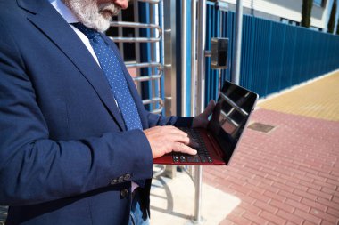 Yetişkin erkek yönetici, gri saçlı, sakallı, güneş gözlüklü, ceketli ve kravatlı sokakta dizüstü bilgisayarına danışıyor. Kavramsal işadamı ve yönetici.