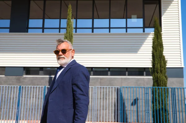 Handsome, mature, grey-haired man with beard, jacket and tie and sunglasses is on the university campus where he works as a university lecturer. Concept professor, university rector.