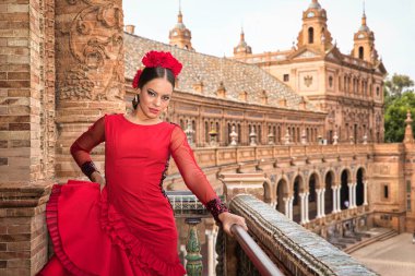 Beautiful teenage woman dancing flamenco on the balcony of a square in Seville. She wears a red dress with ruffles with a lot of art. Flamenco cultural heritage of humanity.