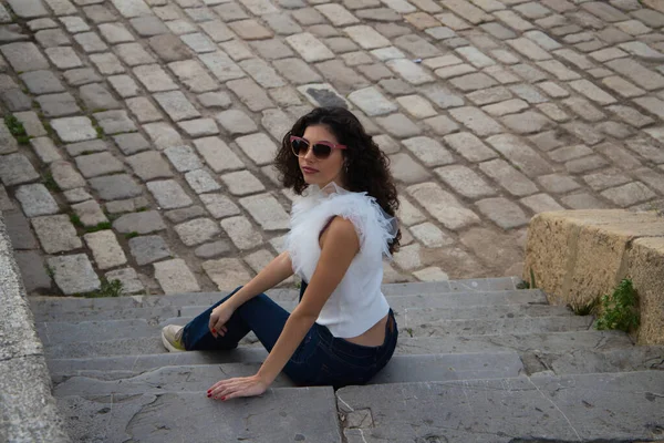 Parktaki bir merdivende oturan siyah, kıvırcık saçlı güzel bir genç kadın. Kadın güneş gözlüğü takıyor ve farklı ifadeler takıyor. İfadeler ve yaşam tarzı konsepti