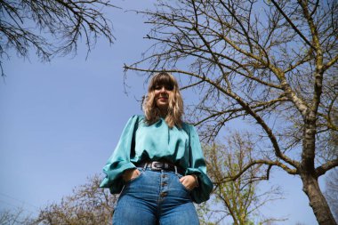 Genç, güzel sarışın kadın yukarıdan kameraya bakıyor, arka planda güneşli bir günde mavi gökyüzünü ve ağaçların dallarını görebilirsiniz. Kadın çok mutlu. İfadeleri kavra