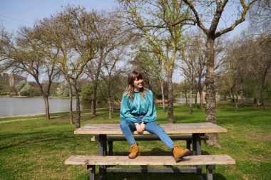 Kot pantolon ve bot giymiş genç, güzel sarışın kadın parktaki ahşap bir masada oturuyor. Arka planda güneşli bir günde nehri görebilirsiniz. Kadın çok mutlu. Tatil kavramı ve ücretsiz.