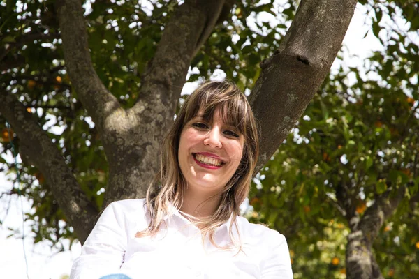 Güzel, sarışın, genç bir kadın kameraya mutlu bir şekilde bakarken bir ağacın tepesinde oturuyor. Kadın, şehirde tatilde. Seyahat ve tatil konsepti.