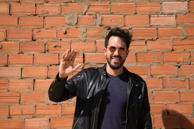 Genç Latin bir adam, esmer, sakallı ve el kaldırarak vücut bulmuş. Arka planda kırmızı tuğla duvar var. Merhaba de, güle güle, selamla. İnsan ilişkileri ve duyguları.